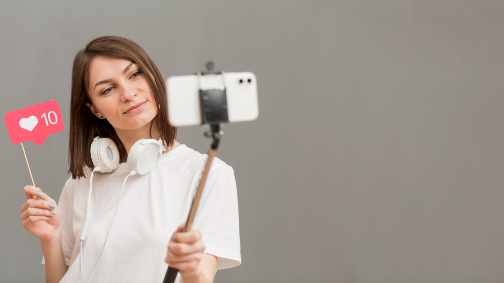 Criadora de conteúdo gravando Reels Virais com smartphone e bastão de selfie, segurando plaquinha de curtidas.